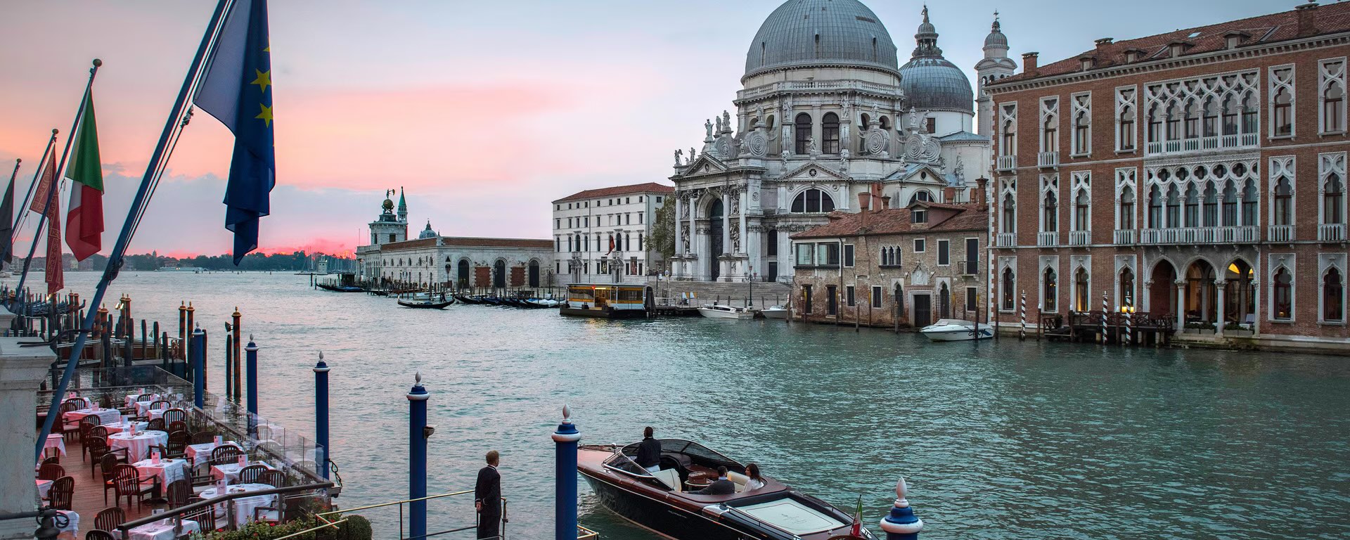 The Grand Canal at dusk seen from the terrace of the Gritti Palace, with laid dining tables and flags in the foreground, a private launch on the water, and the domed basilica of Santa Maria della Salute across the canal.